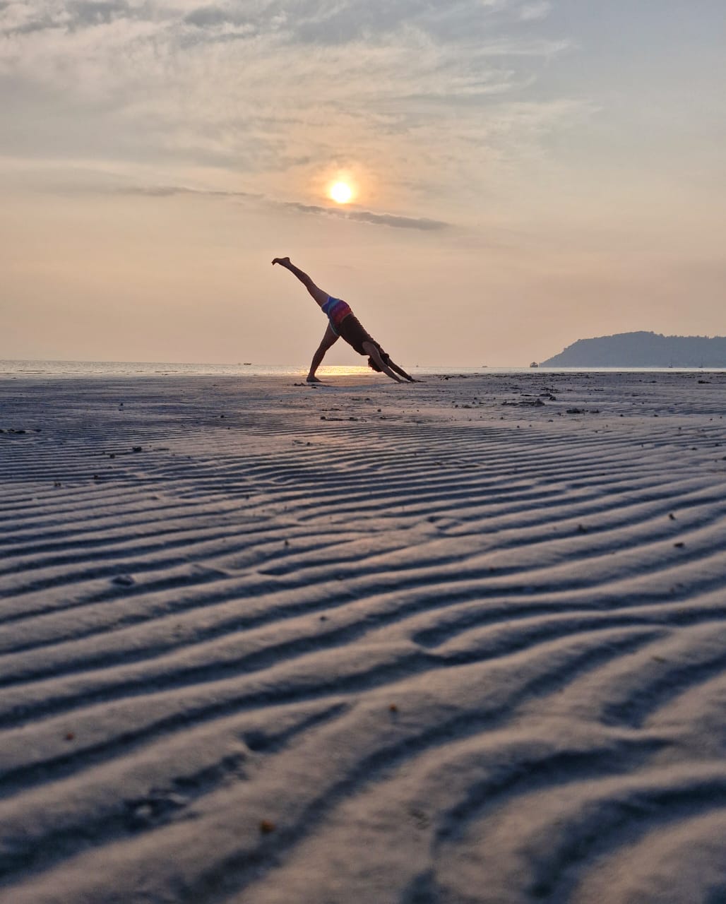 Woman practicing yoga on a rippled beach at sunset, holding a one-legged downward-facing pose by the sea.